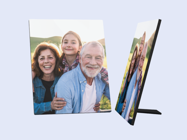 Ein Fotoaufsteller zeigt ein fröhliches Familienbild. Eine lächelnde ältere Frau mit braunen Haaren steht links. Hinter ihr steht ein junges Mädchen mit blonden Haaren in einem karierten Hemd und lächelt ebenfalls. Rechts von ihnen steht ein älterer Mann mit weißen Haaren und Bart, der auch fröhlich in die Kamera schaut und seine Arme um die beiden anderen gelegt hat.